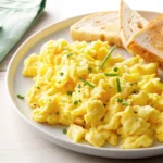 A plate of creamy scrambled eggs with cottage cheese, garnished with chives and served with toast.